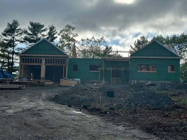 A house under construction with a partially built wooden frame and green siding, surrounded by construction materials and piles of dirt, with a backdrop of trees and a cloudy sky.