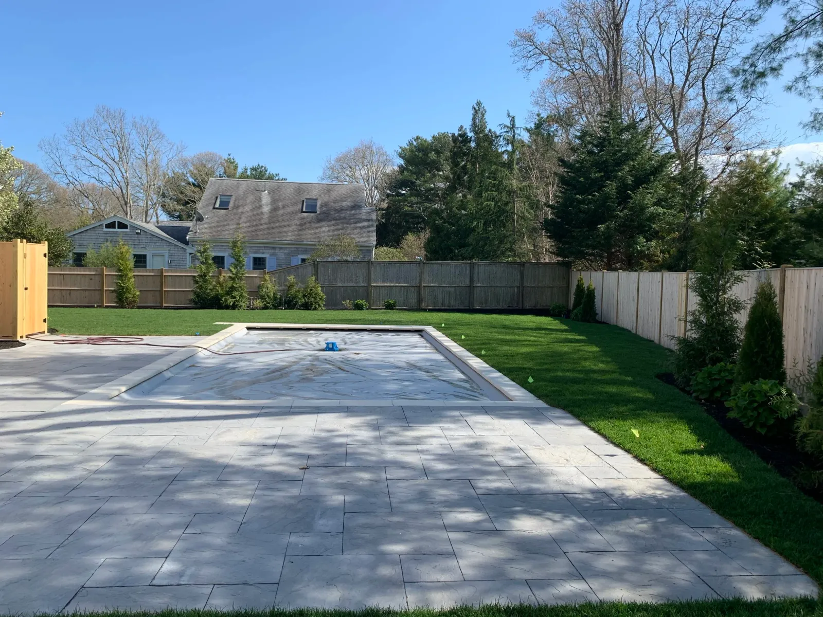 Backyard view featuring a covered swimming pool surrounded by a stone patio. The area has green grass, a wooden fence, and some trees and shrubs. A small house is visible in the background.
