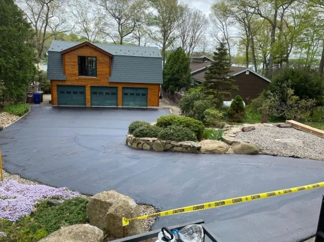 Freshly paved black asphalt driveway in front of a wooden garage with three doors, surrounded by landscaped garden with shrubs and rocks, yellow caution tape across the front.
