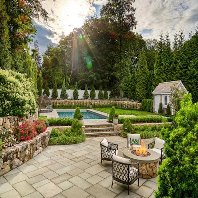 Sunlit backyard with a rectangular pool, stone patio, and a circular fire pit seating area surrounded by lush greenery and trees.