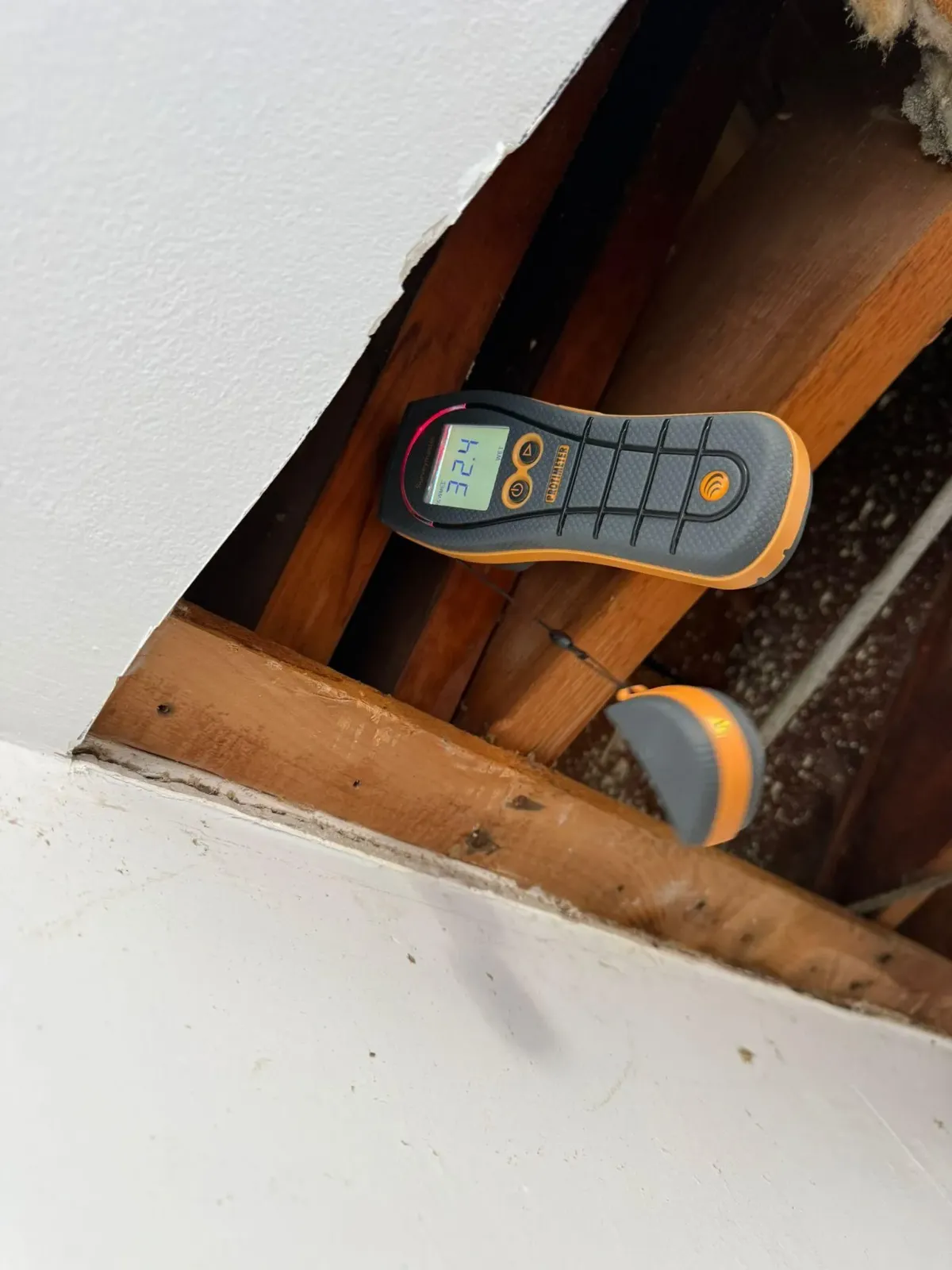 Handheld clamp meter resting on wooden ceiling beams inside an open attic access panel