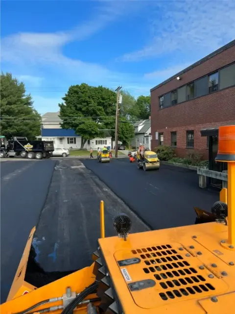 Asphalt paving in progress on a street. Black asphalt, yellow machinery, workers, and a building on a blue sky day.