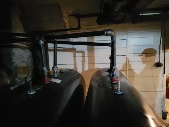 Two black industrial tanks connected by black metal piping in a dimly lit basement.