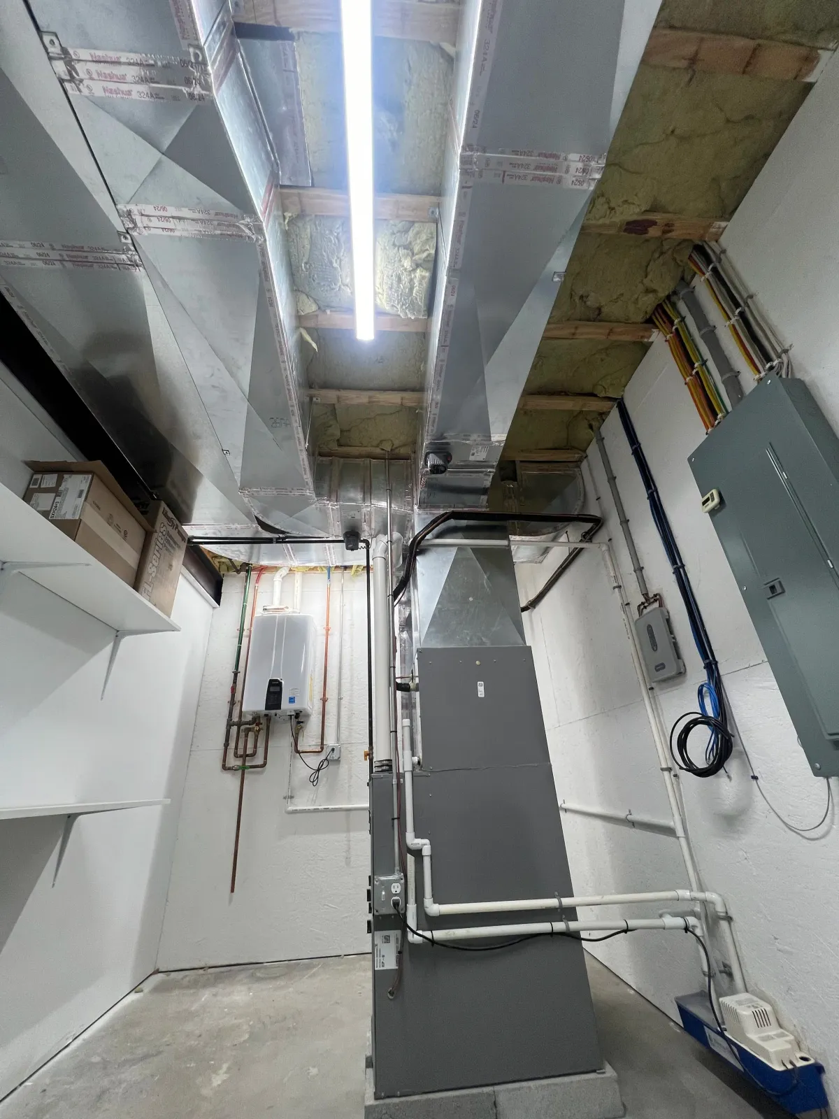 A utility or mechanical room with ductwork, electrical conduits, and pipes installed along the walls and ceiling, with insulation visible behind the ductwork and a fluorescent light fixture overhead.
