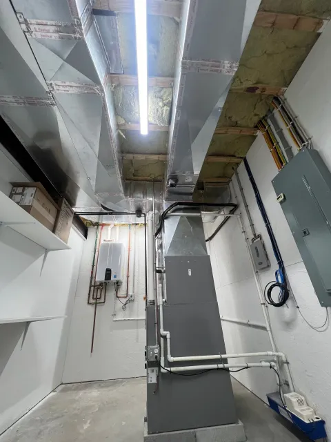 A utility or mechanical room with ductwork, electrical conduits, and pipes installed along the walls and ceiling, with insulation visible behind the ductwork and a fluorescent light fixture overhead.