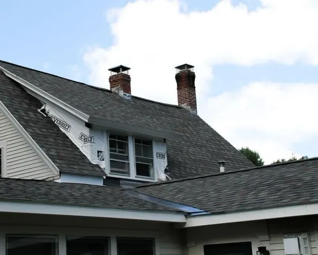 House roof with chimneys, some sections covered in roofing material, others showing underlayment, and windows.