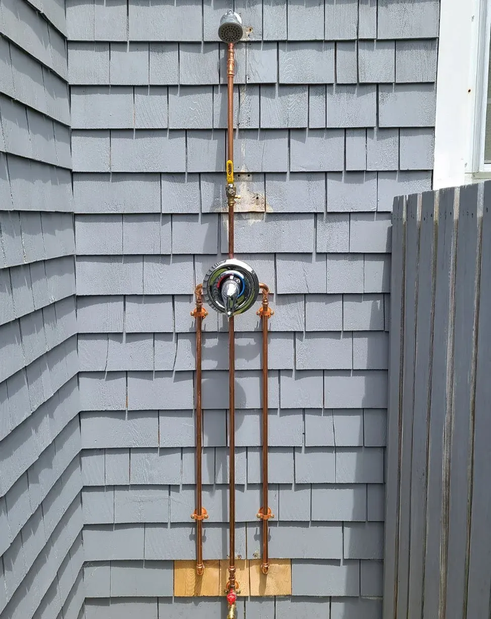 Close-up of outdoor plumbing system on house exterior, featuring copper pipes, a sprinkler head, a valve, and a round, chrome water meter with a dial, mounted on a gray shingled wall.