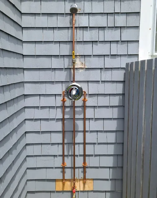 Close-up of outdoor plumbing system on house exterior, featuring copper pipes, a sprinkler head, a valve, and a round, chrome water meter with a dial, mounted on a gray shingled wall.