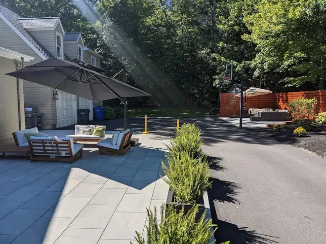 Back patio with lounge chairs, umbrella, potted plants, and sunlit driveway beside a house
