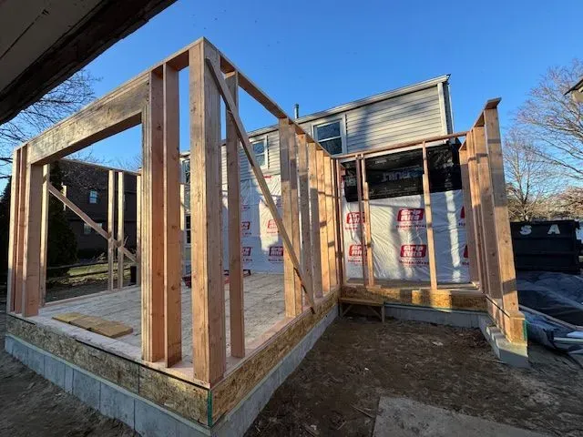 A wooden house addition in the framing stage, featuring unfinished wall studs on a foundation against an existing home.