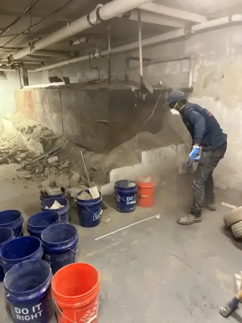 A construction worker removes rubble and debris from a basement area into several plastic buckets near a large metal tank.