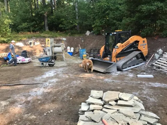 Construction site with a mini excavator, a person, a dog, and materials like stones, buckets, and pallets surrounded by trees.