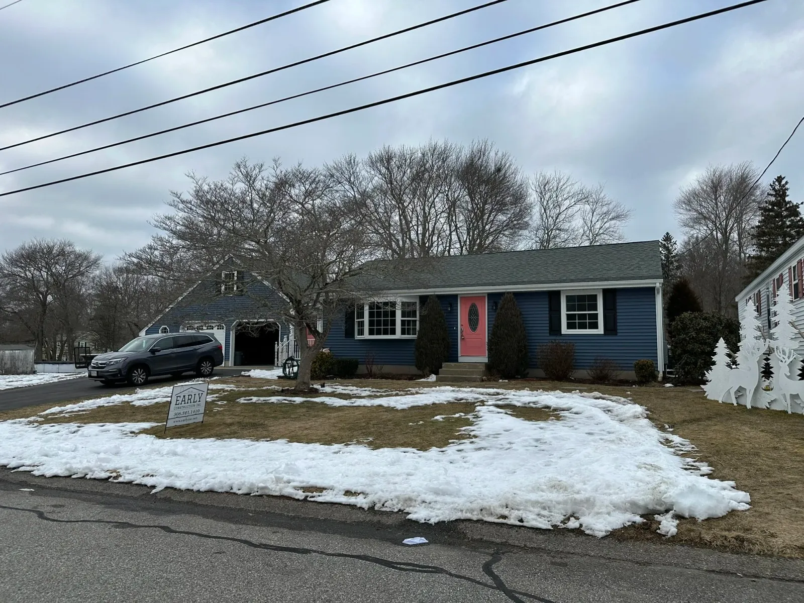 A blue house with a pink front door, two bushes flanking the door, and a snow-covered lawn. There is a black car parked in the driveway, a sign that reads 'Early,' and a white fence with trees in the background, under a cloudy sky.