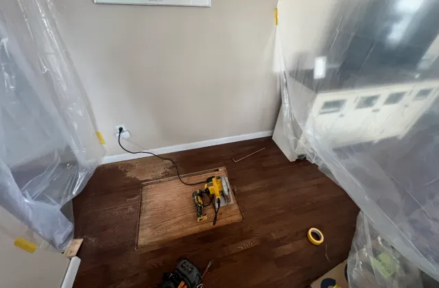 Room with a section of hardwood floor being replaced, a power drill and a chisel on the floor, plastic sheeting covering cabinets and walls, and an electrical outlet with a black cord plugged in.