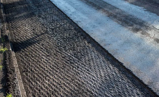A road shoulder showing freshly milled, textured asphalt running parallel to a smooth, existing paved lane.
