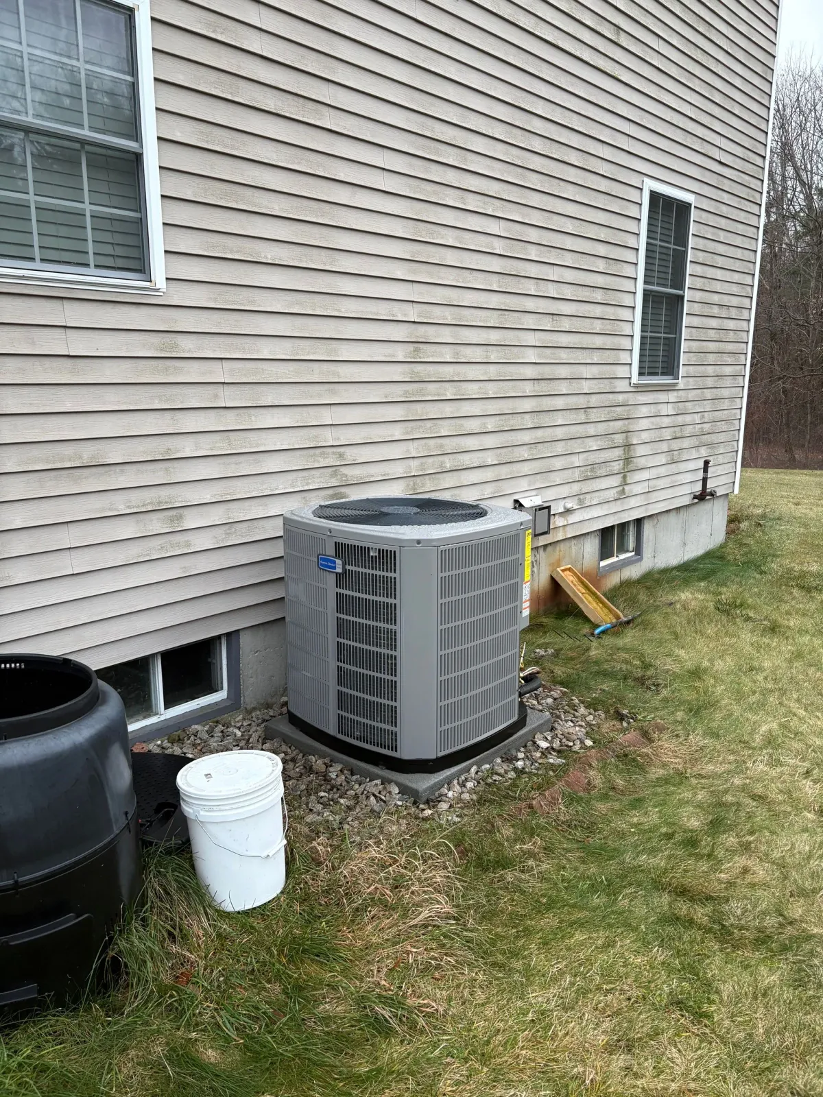 Air conditioning unit next to a light-colored building with a window, surrounded by grass and a composting bin.