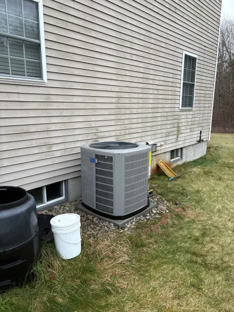 Air conditioning unit next to a light-colored building with a window, surrounded by grass and a composting bin.