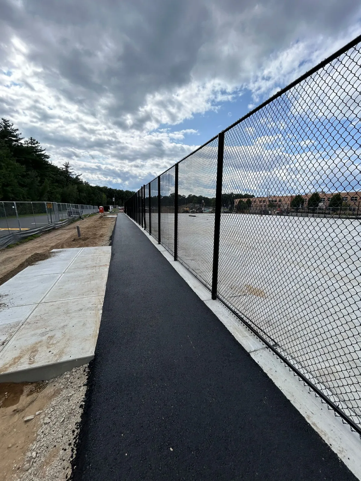 A newly paved black asphalt path runs alongside a tall chain-link fence, with trees and a cloudy sky in the background.