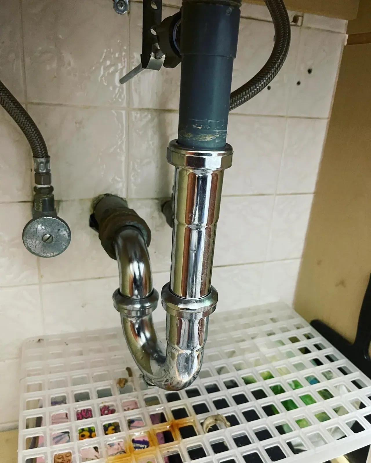 Under kitchen sink plumbing with metal pipes, a black pipe, and a valve with a red handle, against tiled wall, with a plastic grid organizer containing various colored items below.