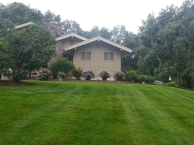 Large green lawn in front of a beige house with trees and shrubs surrounding it.