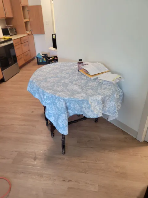 A round table covered with a blue floral tablecloth, with papers and a bottle on top, in a room.