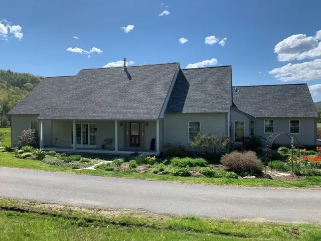 A light gray house with a dark gray roof sits next to a paved driveway under a blue sky.
