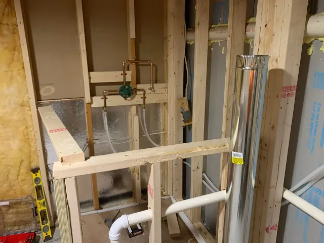Framed walls with pipes, plumbing fixtures, and metal chimney in construction.