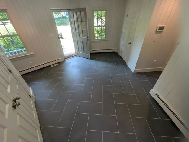 Empty room with dark gray tiled floor, white paneled walls, and open door leading outside to greenery and water. Windows with views of trees and a deck area.