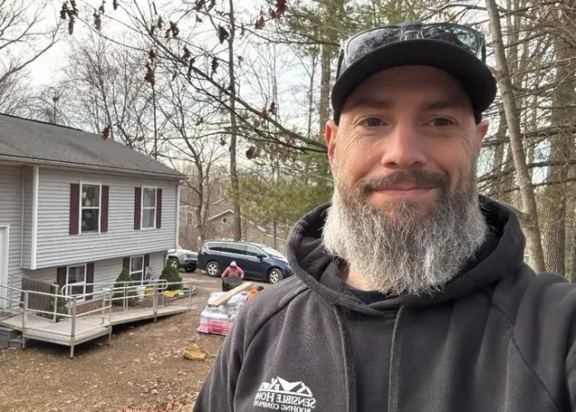Man with grey beard and cap in front of a two-story house with a car and people in the background.