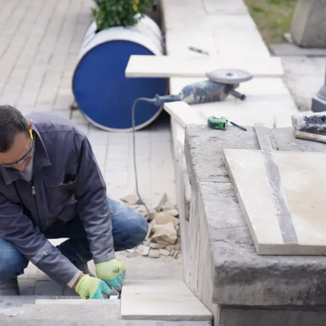 Mason kneeling to fit a pale stone cap onto a low stone wall beside his tools