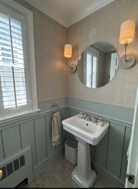A small bathroom with a pedestal sink, a round mirror, two wall-mounted lamps, a window with white shutters, and light green wainscoting on the walls.