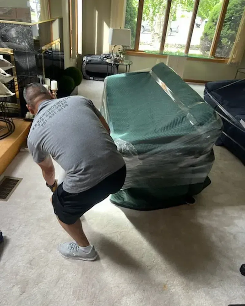 A person in gray shirt and black shorts lifting a large, wrapped object in a room.