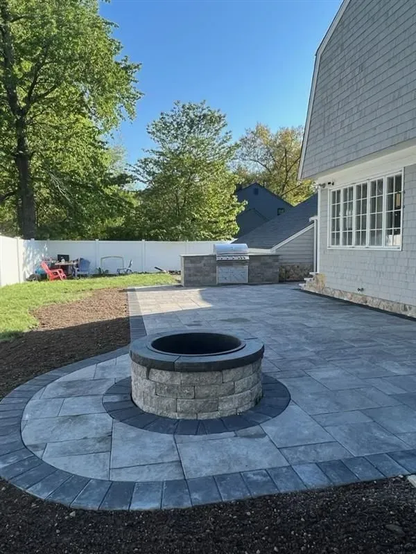 A gray stone patio featuring a circular fire pit, a built-in outdoor grill, and a grassy yard with a white fence.