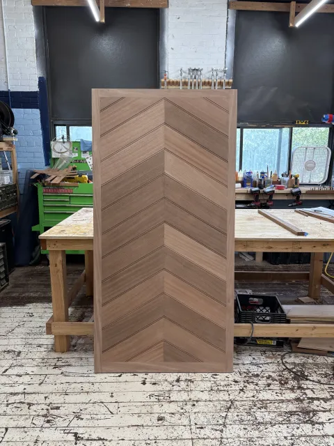 A woodworking workshop with a wooden panel featuring a chevron pattern in the foreground. The workshop has a workbench with tools, cordless drills, and supplies. Shelves hold clamps, and there is a window providing natural light. The floor is worn wo