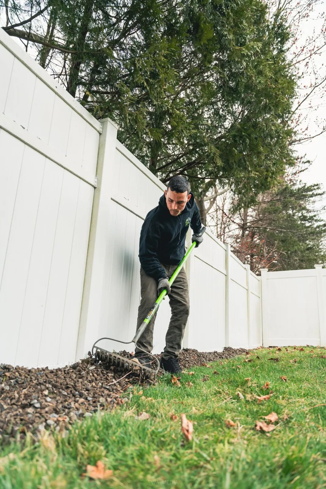 A man using a rake to level small rocks along the edge of a lawn, next to a white fence and tall trees.