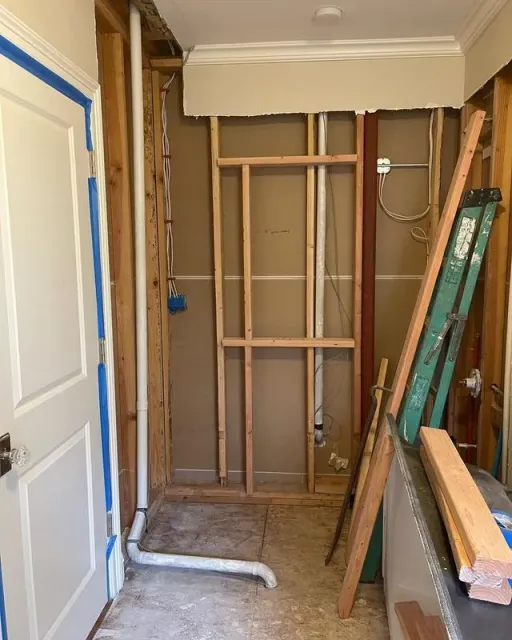 Bathroom under construction: walls open, plumbing visible, new frame, ladder and wood.