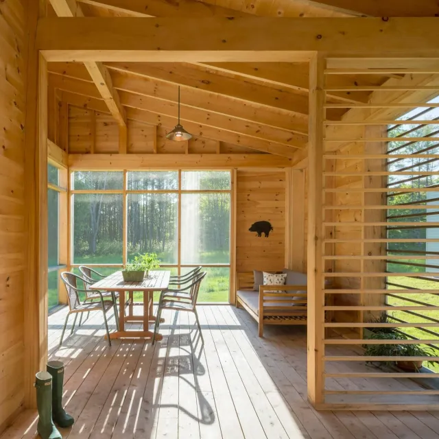 A screened porch with wooden walls, ceiling, and floor, featuring a large window overlooking a green landscape with trees, a wooden dining table with four chairs, a bench with pillows, and a pair of green rain boots near the door.