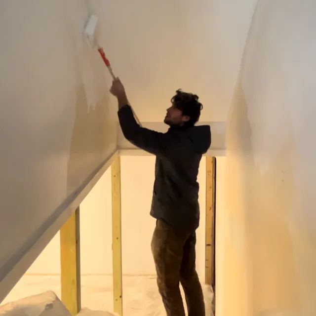 A person painting a ceiling with a roller brush in an unfinished room with exposed wooden framing.