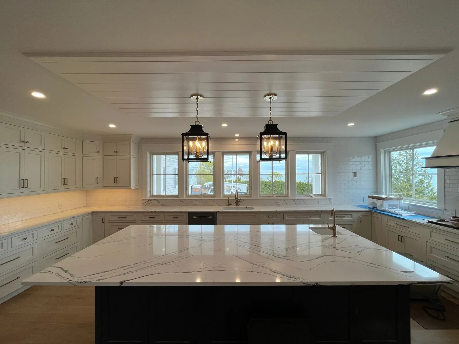 Bright, spacious kitchen with white cabinets, a large marble island, and three pendant lights hanging above the island. Multiple windows provide natural light and a view of trees outside.