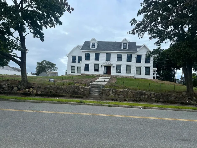 A large white house on a hill with a stone foundation, featuring multiple windows and a front entrance with steps, surrounded by trees and a cloudy sky.