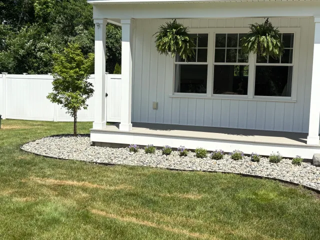 White house exterior with a large window, two hanging ferns, a small tree, and a white fence.