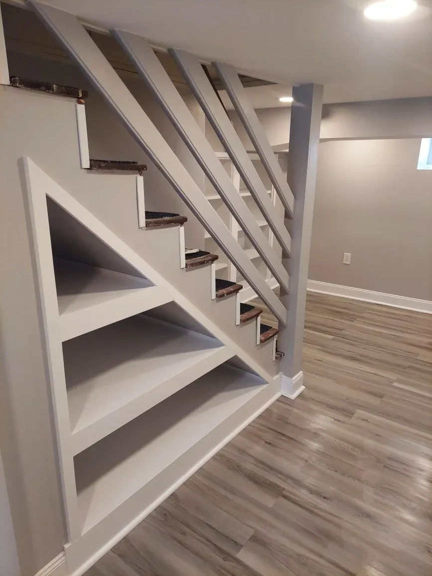 Partially constructed staircase with a bookshelf underneath, white painted walls, wood flooring, and a small window in a modern interior.