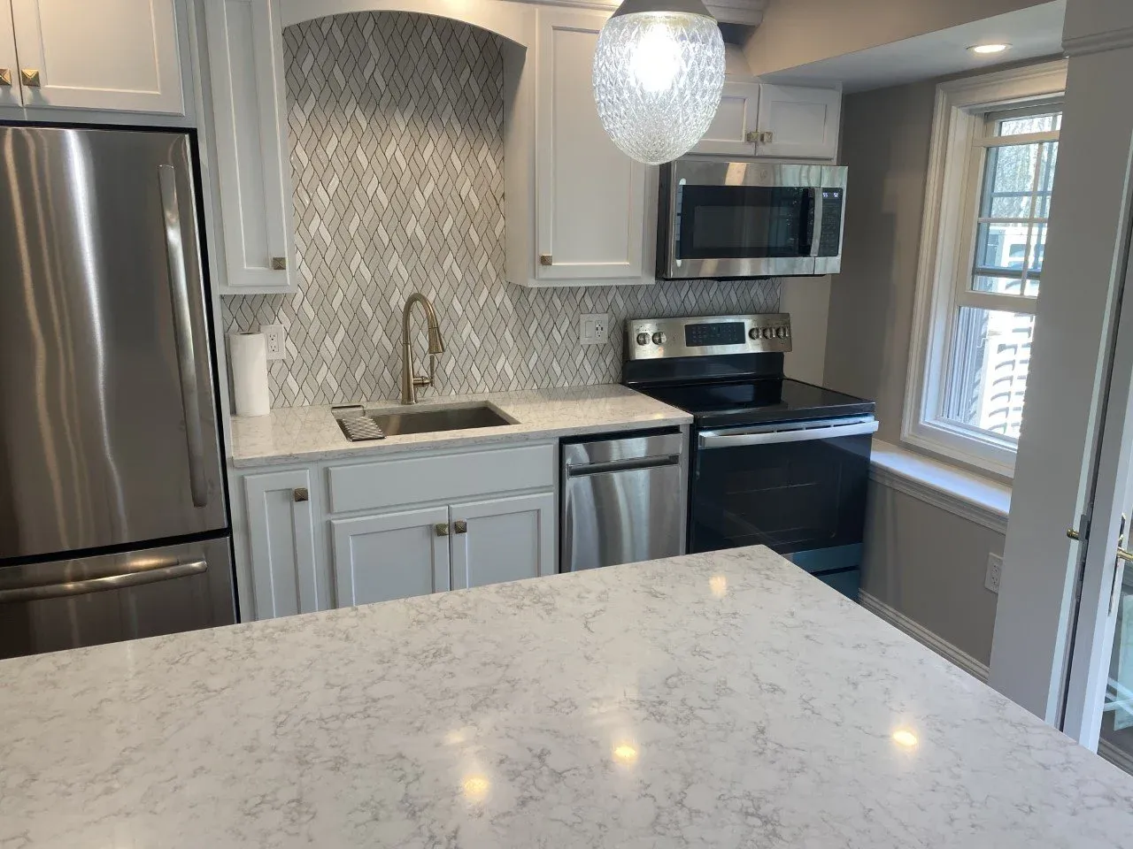 Modern kitchen with white cabinets, marble countertops, stainless steel appliances, a black stove, microwave, and refrigerator, with a geometric backsplash and a large window.