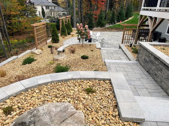A landscaped backyard with stone pathways, decorative gravel areas, wooden trellises, and some trees. The area includes pavers, shrubs, and outdoor furniture on a patio.