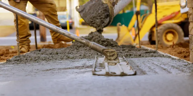 Concrete being poured onto a surface by a construction worker using a shovel, with construction equipment and workers in the background.