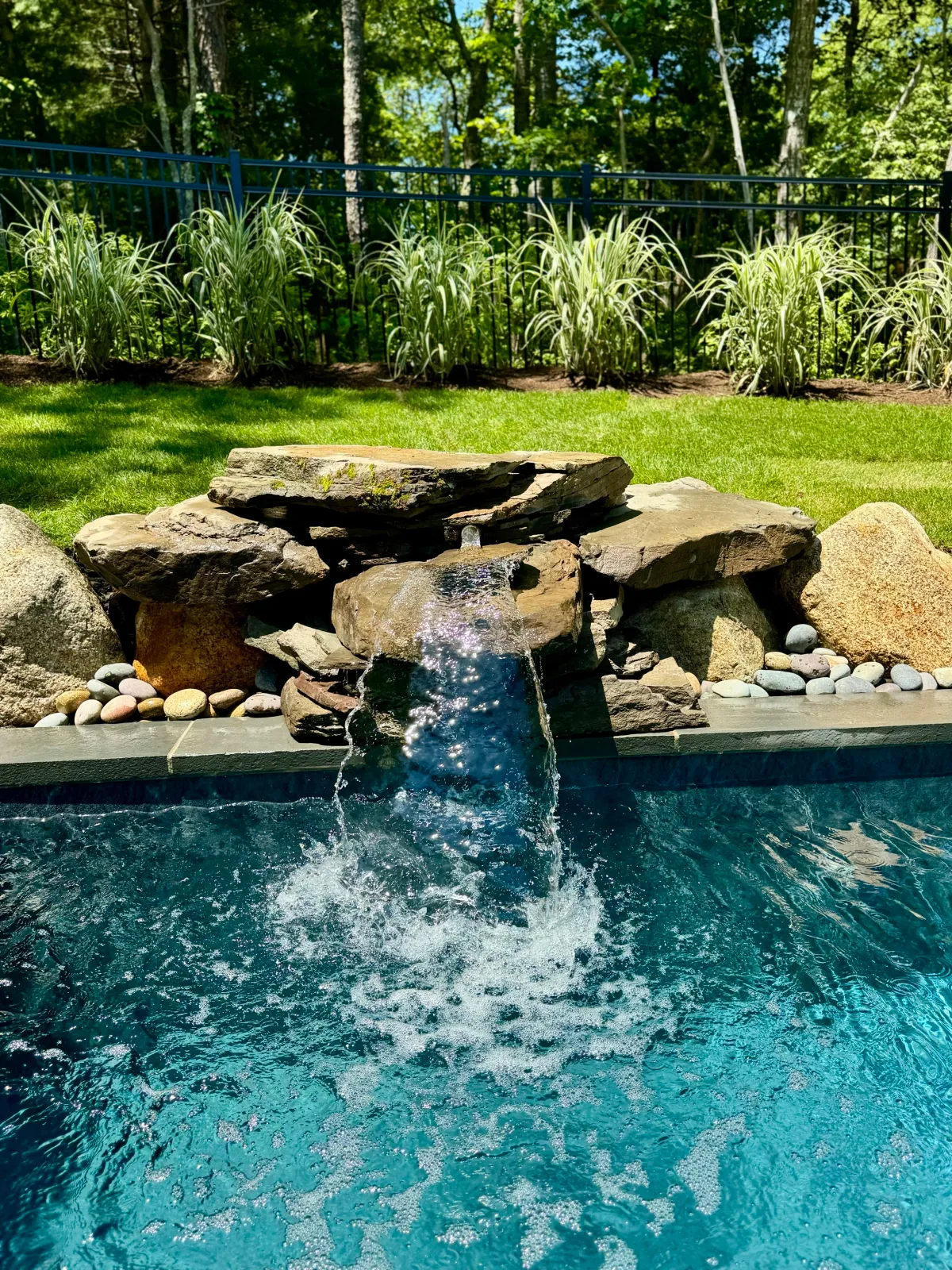 Waterfall feature with stone accents in a pool, surrounded by a grassy lawn and ornamental grass, with a metal fence and trees in the background.