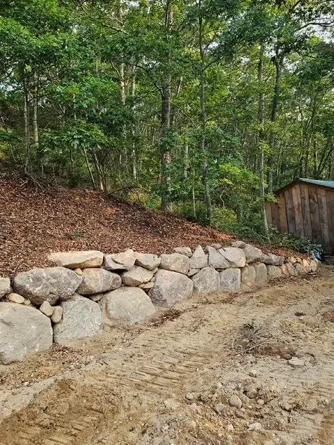 Rock retaining wall along a dirt path beside a wooded hillside and small shed