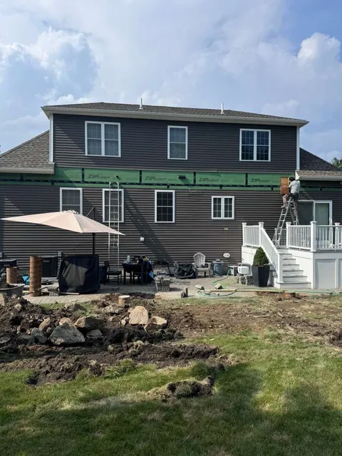 A worker on a ladder installs siding on the back of a two-story gray house with a deck and a messy yard under a blue sky.