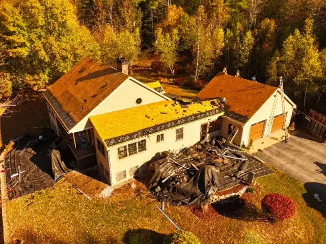 Aerial view of a house with damaged roofs covered in debris, surrounded by autumn trees and construction materials on the ground.