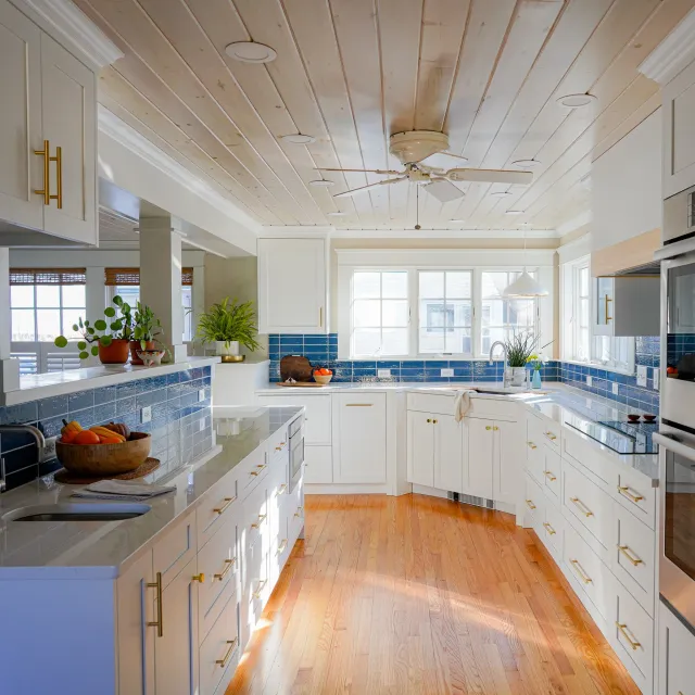 Bright white kitchen with blue tiled backsplash, white cabinets with gold handles, wooden floors, and large windows letting in natural light.